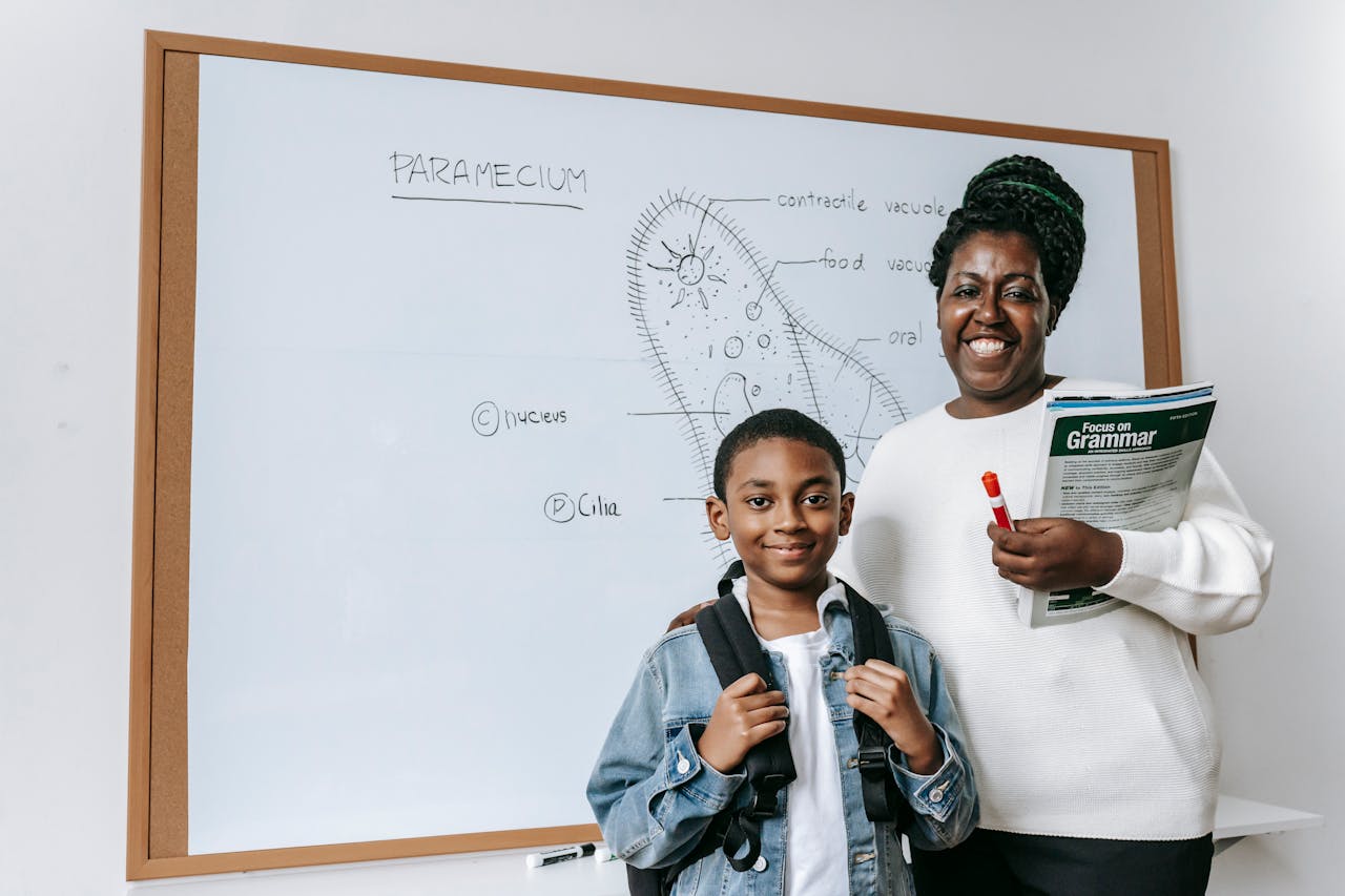 Smiling female teacher holding textbooks and standing near African American boy with backpack while looking at camera